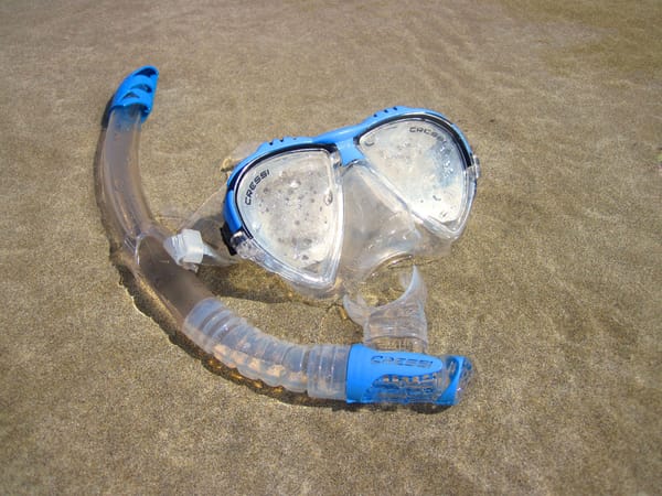 A snorkel and a dive mask in very shallow water, atop some beach sand.